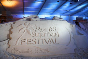 A sand sculpture with carvings of a palm tree scene and the words "Pier 60 Sugar Sand Festival."